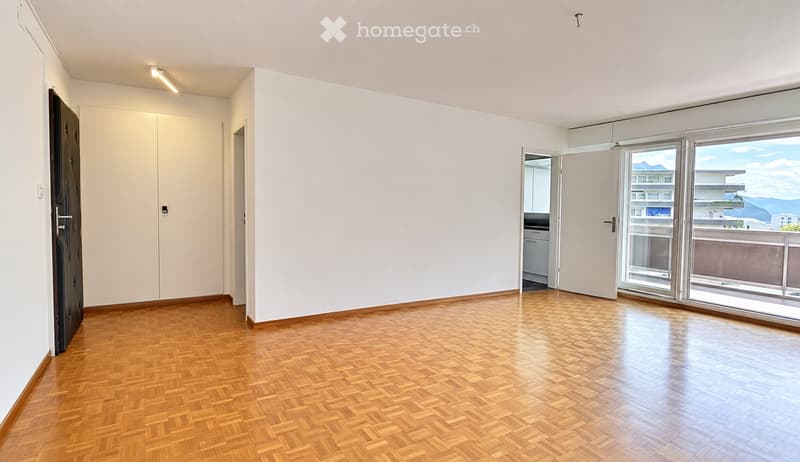Empty room with wooden herringbone flooring, white walls, large windows, and a door leading to a balcony.