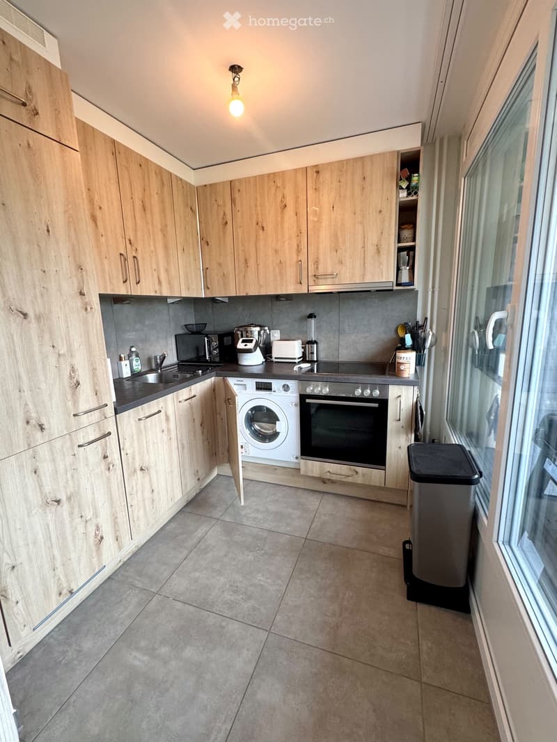Modern kitchen with light wooden cabinets, stainless steel appliances, and a tiled floor.