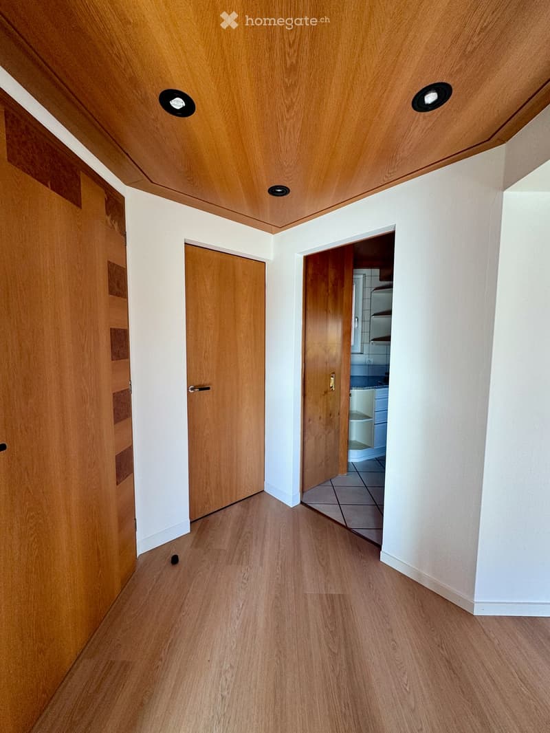 A clean, well-lit hallway with wooden flooring, white walls, and recessed ceiling lights. Two wooden doors are visible, one leading to a room and the other to a bathroom.