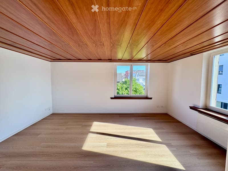 Empty room with wooden flooring, white walls, and large windows allowing natural light to enter. The ceiling features a wooden panel design.