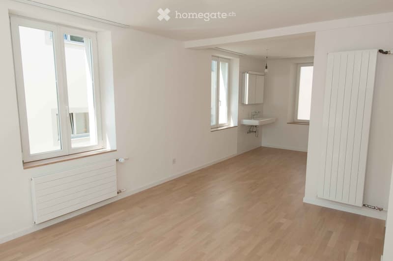Empty room with light wooden flooring, white walls, and large windows. A small adjacent bathroom area is visible with a sink and mirror.