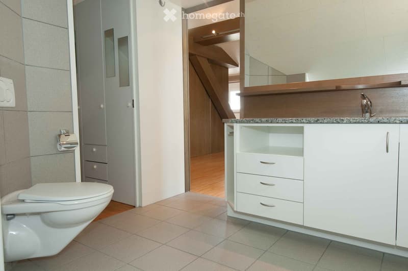 Modern bathroom with a white toilet, a white sink with a granite countertop, a mirror, and a white cabinet with drawers. The floor is tiled, and the walls are tiled with a grey finish. A wooden staircase is visible in the background.