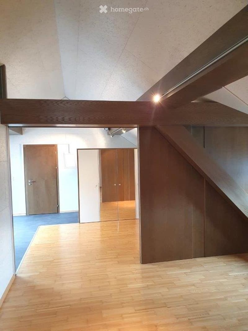 Empty room with wooden flooring, white walls, and a wooden door visible in the background. The space is well-lit with natural light coming from the ceiling.