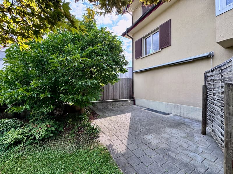 Garden area with paved stone pathway, greenery, and wooden fence adjacent to a building with windows and shutters.