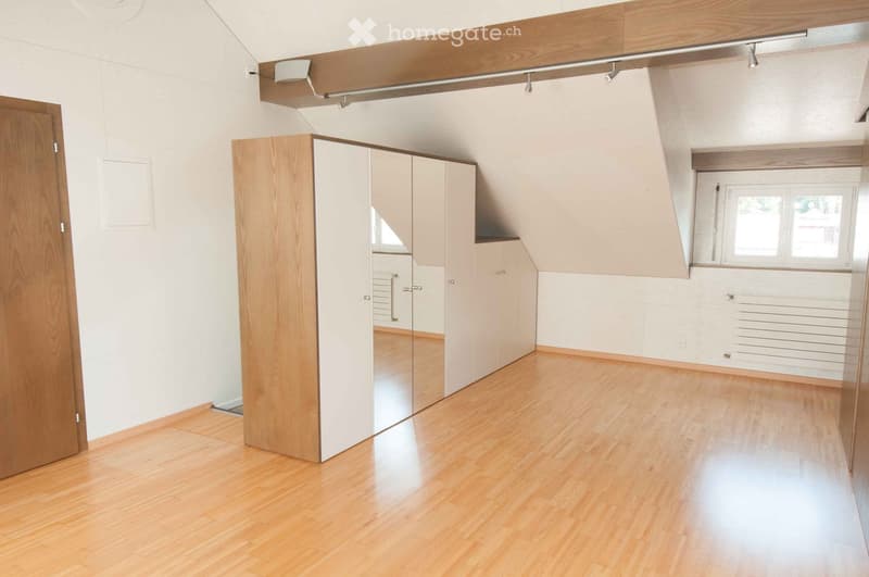 Empty room with wooden flooring, white walls, built-in closet with mirrored doors, and a window letting in natural light.