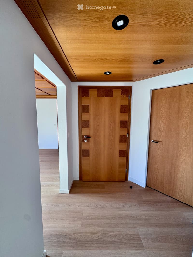 Empty hallway with wooden flooring, white walls, and two wooden doors. The ceiling features recessed lighting and a wooden panel design.