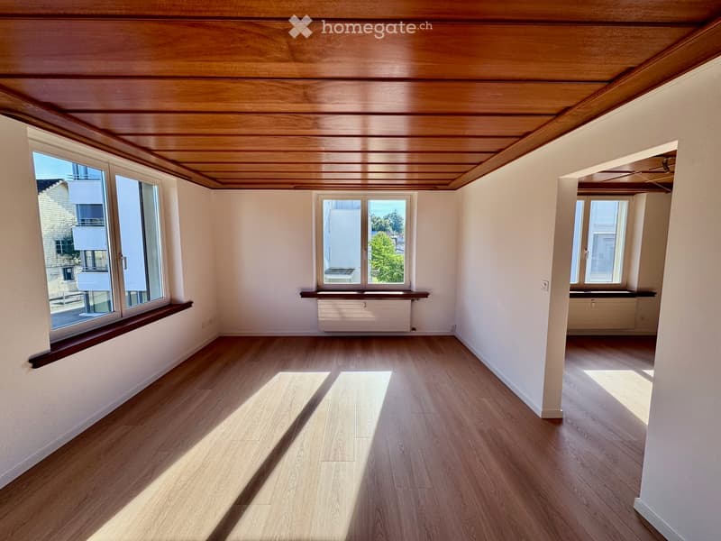Empty room with wooden flooring, white walls, and large windows allowing natural light. The room features a wooden ceiling and built-in window sills.
