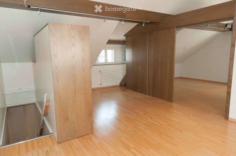 Empty room with wooden flooring, white walls, and exposed wooden beams on the ceiling. The room features a large window with blinds and a radiator below it.