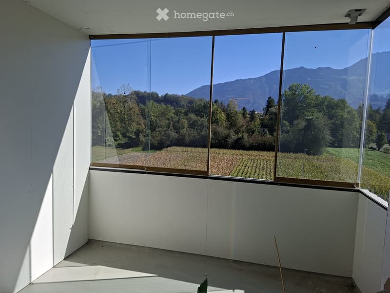 Empty room with large windows offering a scenic view of mountains and fields. Bare walls and floor, no furniture.