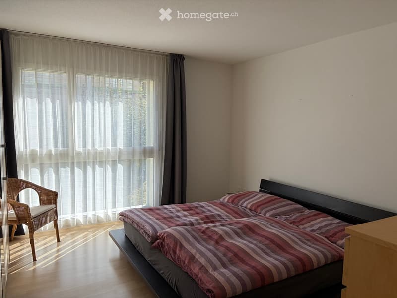 Bright bedroom with wooden flooring, large window with sheer curtains, double bed with striped bedding, and a wicker chair near the window.