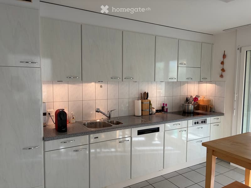 Modern kitchen with white cabinets, granite countertops, stainless steel sink, dishwasher, coffee maker, and wooden table.