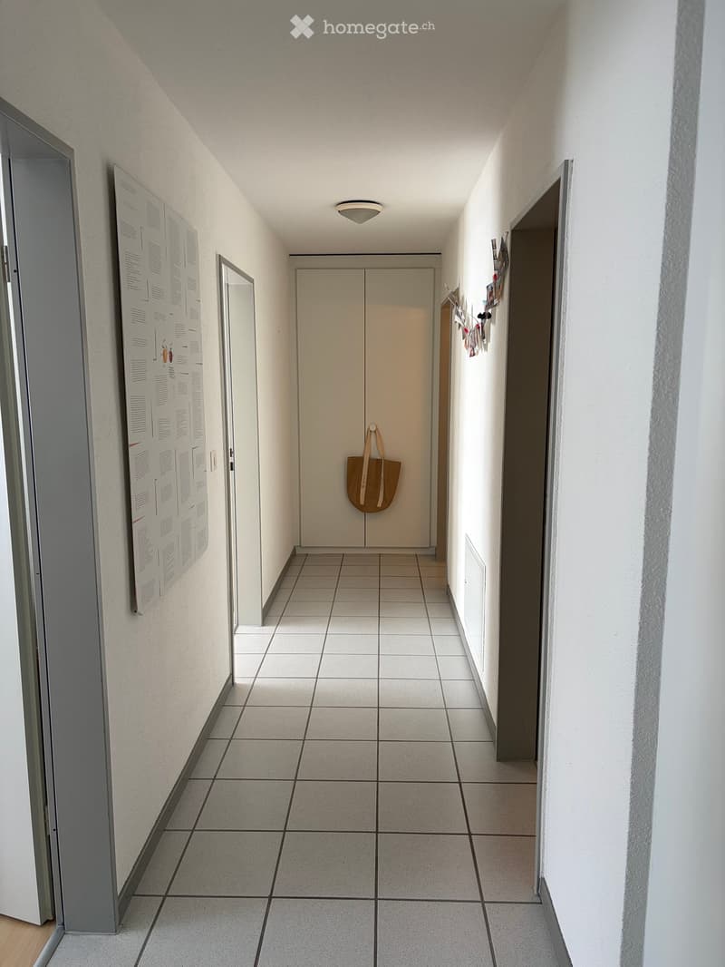 A narrow hallway with tiled flooring, white walls, and a single overhead light fixture. A closed door is visible at the end, with a small bag hanging on it. A bulletin board is mounted on the left wall, and a mirror is visible on the left side.