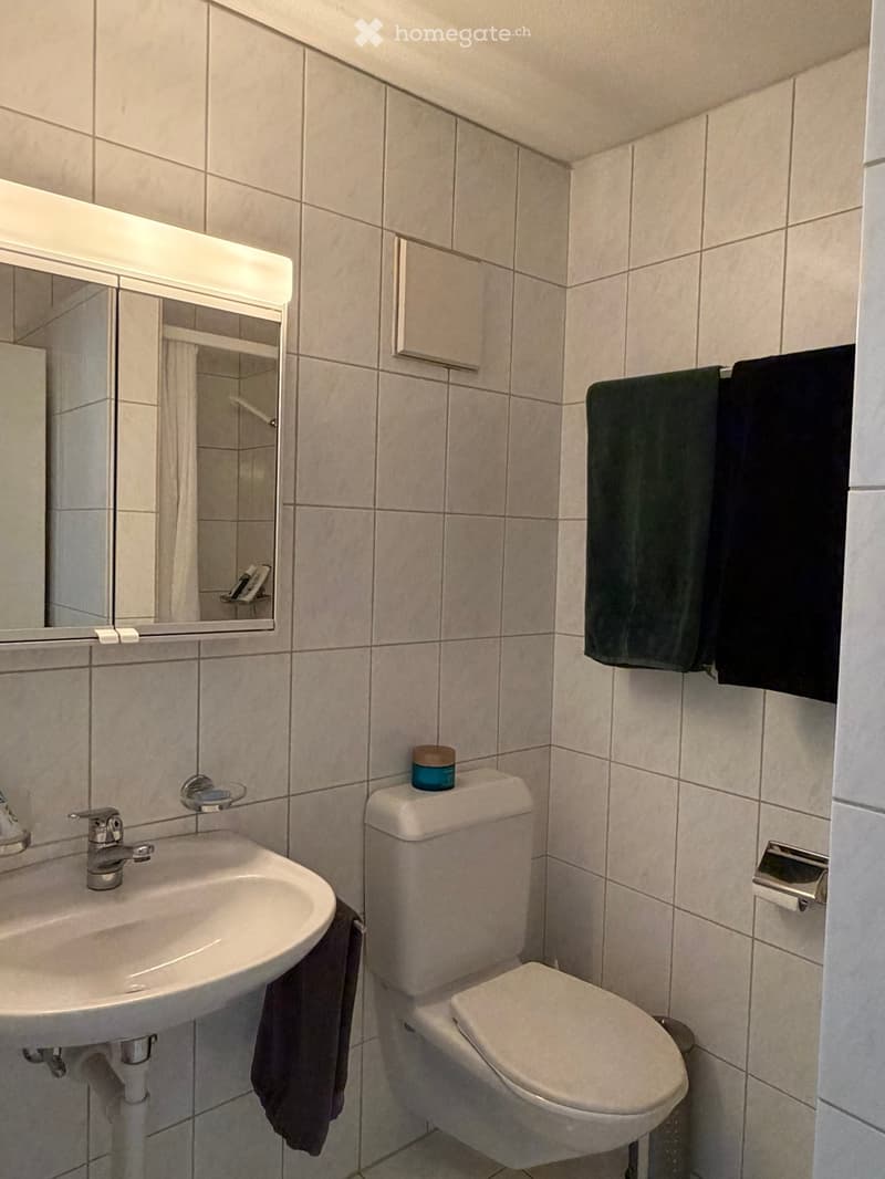A small bathroom with a white tiled wall and floor, featuring a sink with a silver faucet, a toilet, a mirror with built-in lighting, and towels hanging on a rack.