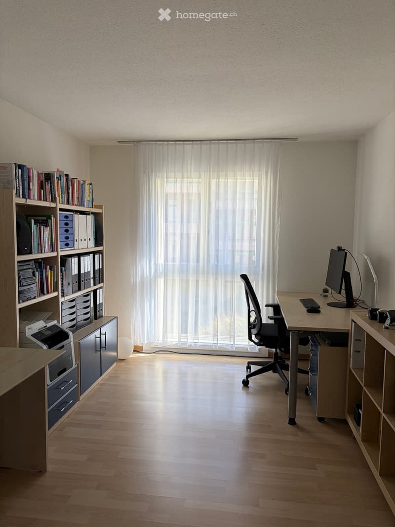 A well-lit office space with a wooden floor, a large window with sheer curtains, a desk with a computer setup, a bookshelf filled with books and office supplies, and a comfortable office chair.