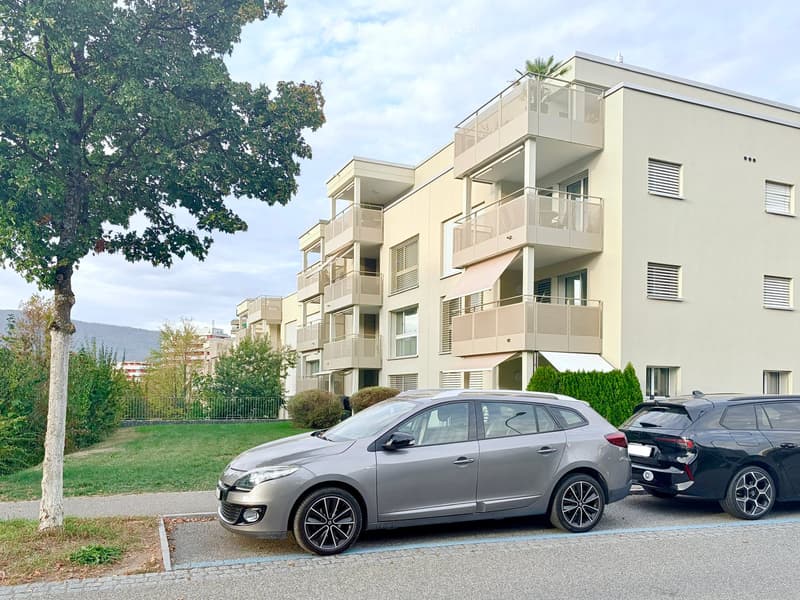 Modern multi-story apartment building with balconies, parked cars in front, surrounded by greenery and trees.