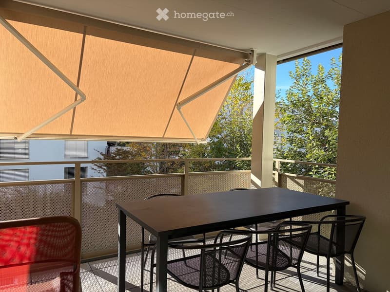 Balcony with a retractable awning, a black table, and four black chairs, overlooking a tree-lined area.