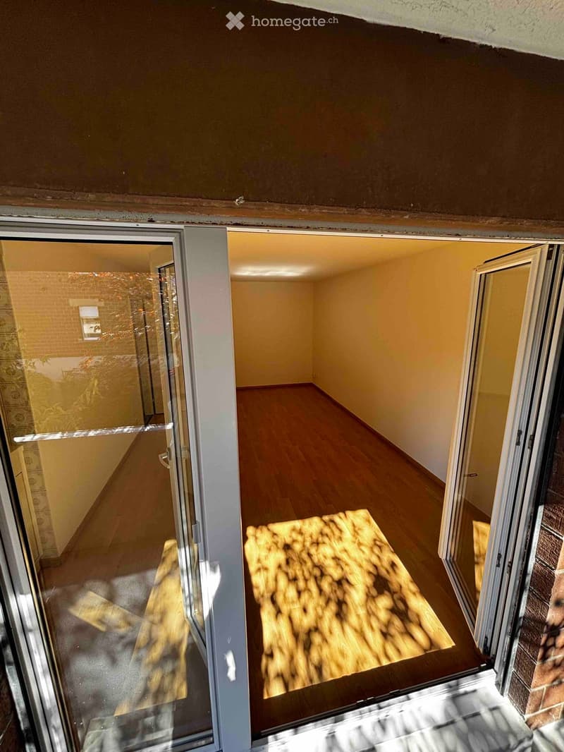 Empty room with wooden flooring and light yellow walls, visible through an open sliding glass door.