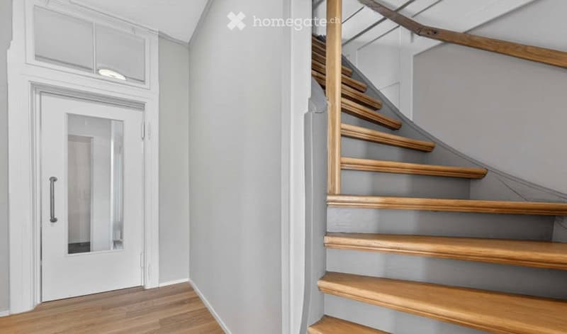 A modern hallway with a wooden staircase featuring light grey walls and a white door with a glass panel.