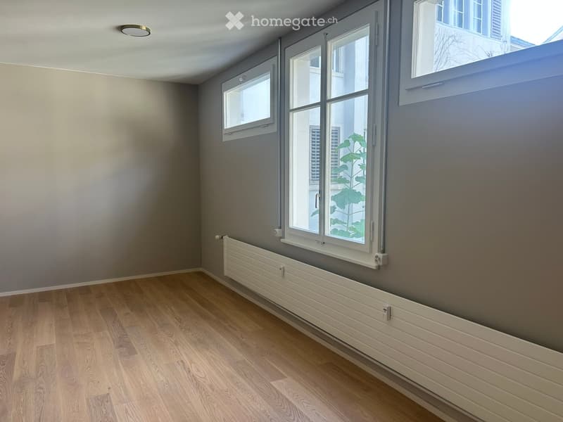Empty room with wooden flooring, grey walls, and multiple windows allowing natural light. A radiator is visible under the windows.