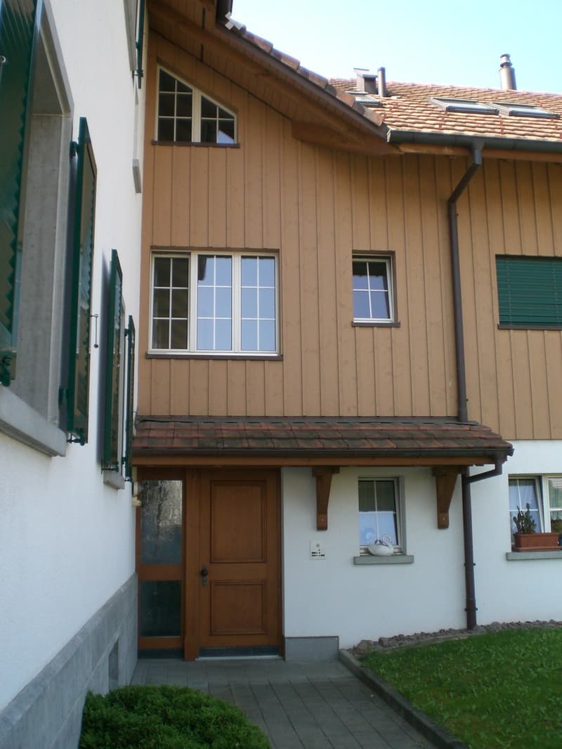Two-story house with wooden siding, multiple windows, and a tiled roof. Features a wooden front door with glass panels, a small garden area, and decorative items on the windowsills.