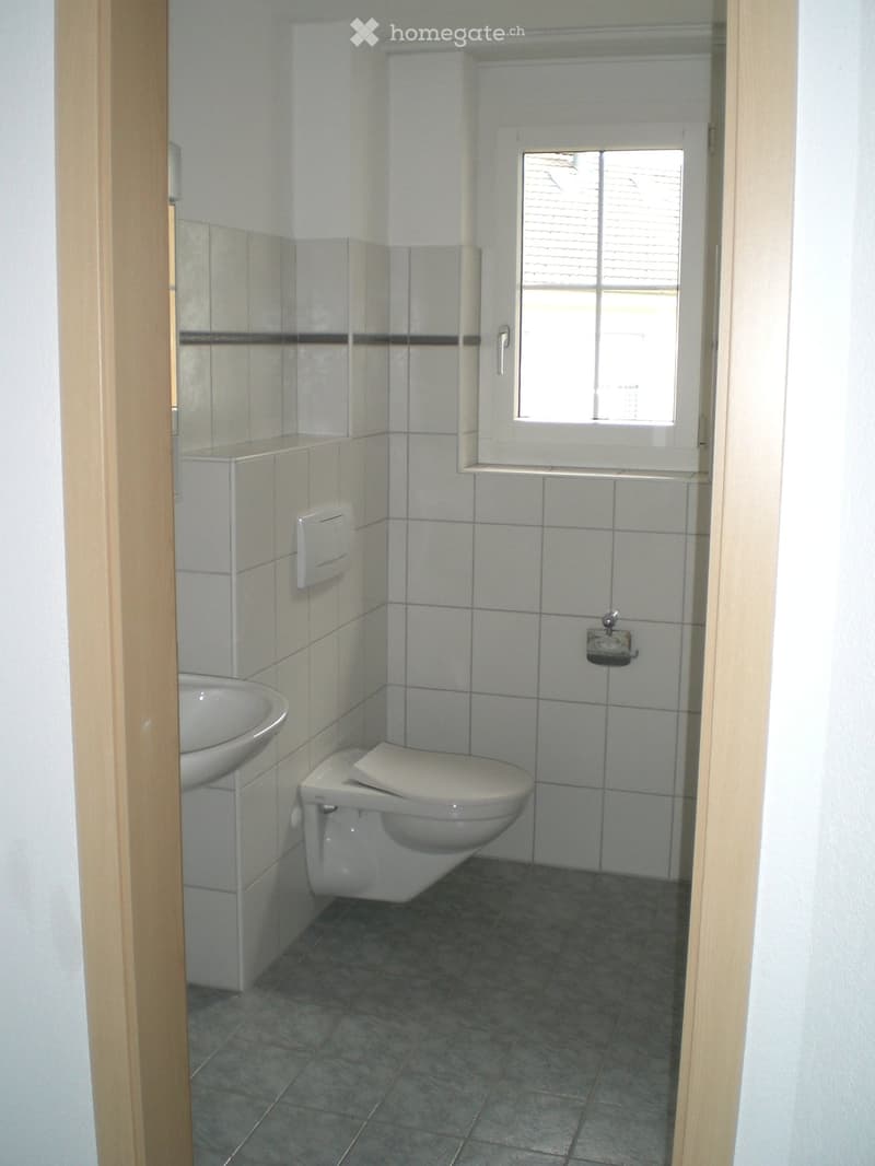 Small bathroom with white tiled walls, a toilet, a sink, and a window with blinds.