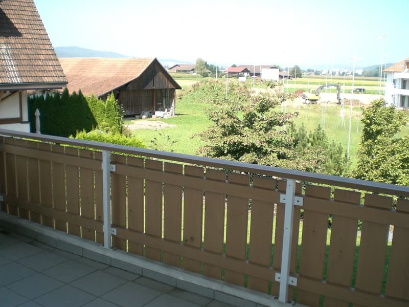 Balcony with wooden railing overlooking a green lawn, trees, and distant houses.