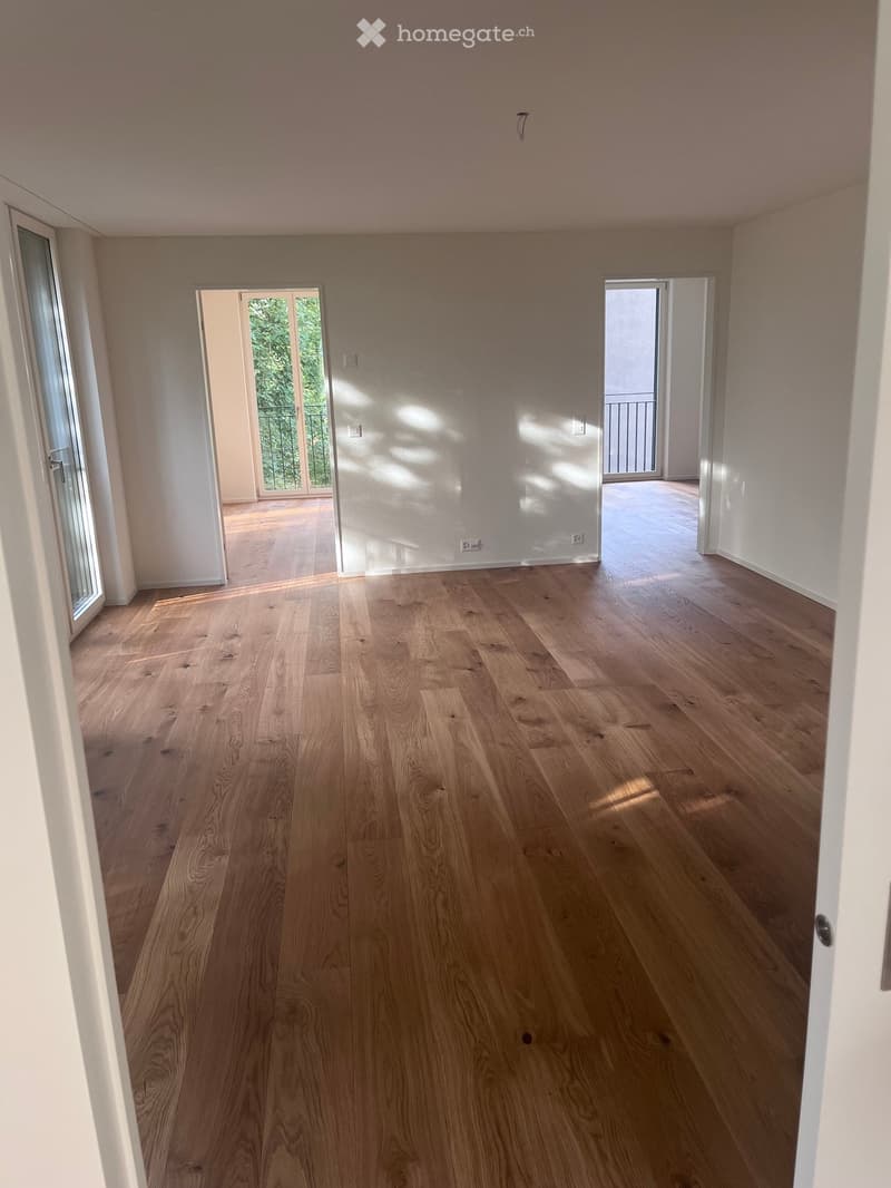 Empty room with light wooden flooring, white walls, and large windows allowing natural light. The room has two open doorways leading to other spaces.