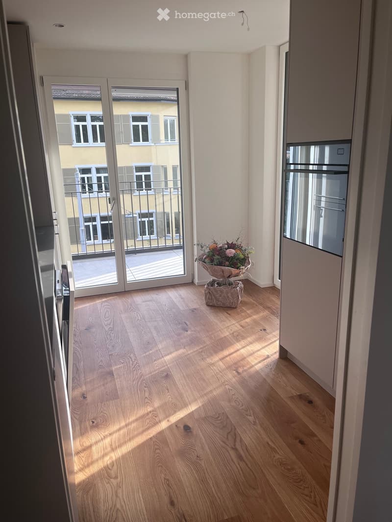 Empty hallway with wooden flooring, large sliding glass doors leading to a balcony, and a small flower arrangement on the floor.