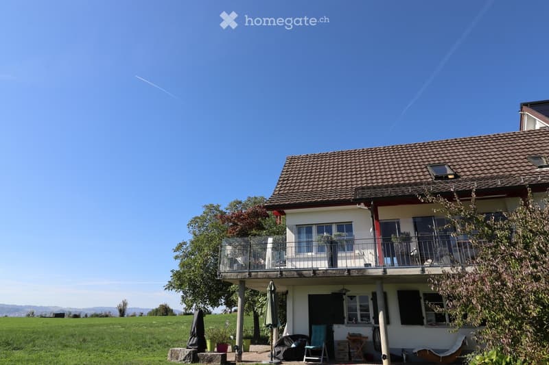 Two-story house with a tiled roof, balcony, and surrounding garden area with outdoor furniture.