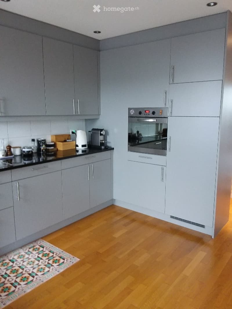 Modern kitchen with grey cabinets, wooden flooring, and various appliances including an oven, coffee maker, and cups on the counter.