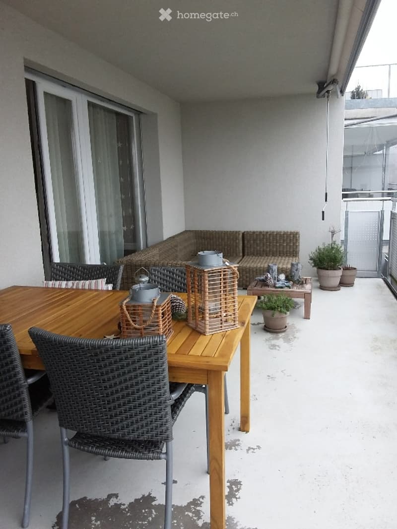 Balcony with wooden dining table, wicker chairs, sofa, lanterns, and potted plants.