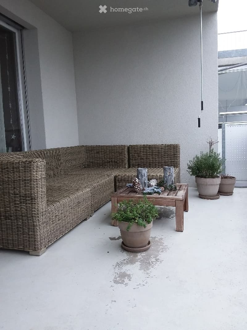 A modern, minimalist balcony with a wicker sofa, wooden coffee table, and several potted plants. The space features clean lines and neutral tones, with a glass door leading to the interior.