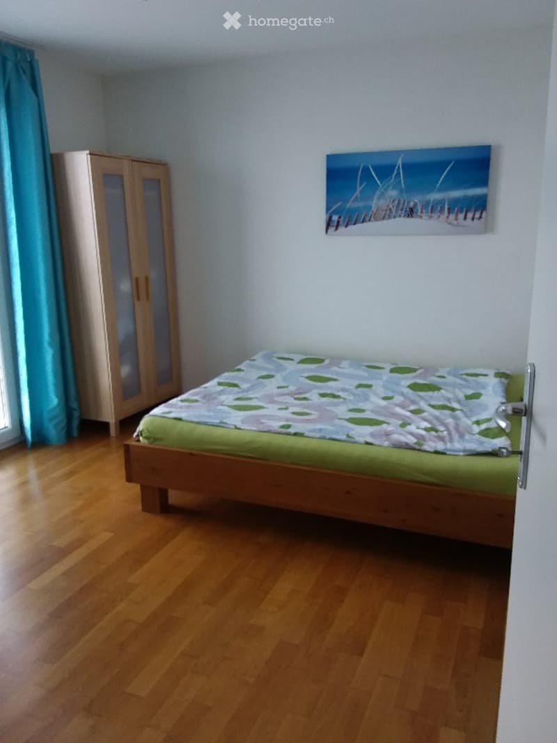 Bedroom with wooden floor, single bed with green and white bedding, light-colored wardrobe, and a painting on the wall.