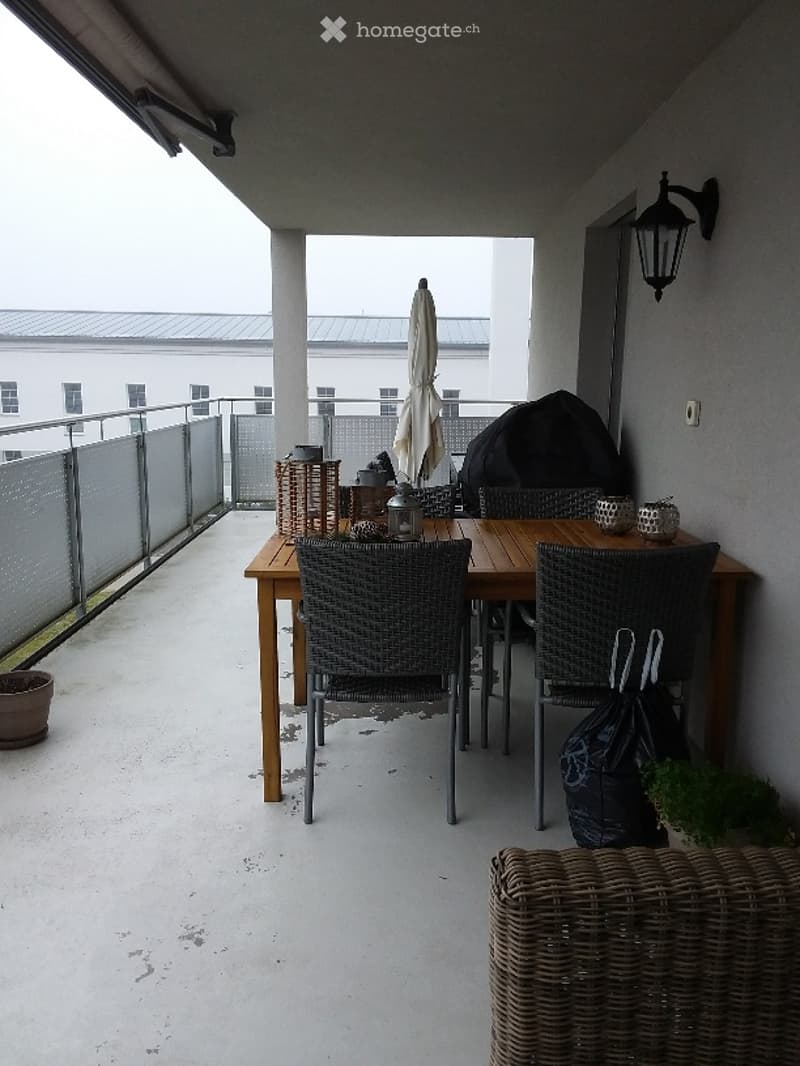 Balcony with a wooden dining table, wicker chairs, and a closed umbrella. The balcony has a metal railing and a view of a building in the background.