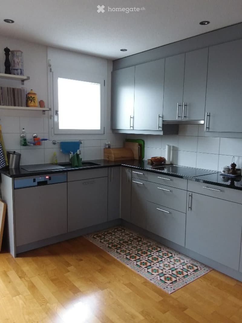 Modern kitchen with grey cabinets, black countertops, and wooden flooring. Features include a sink, dishwasher, stove, oven, and various kitchen items on the counters and shelves.