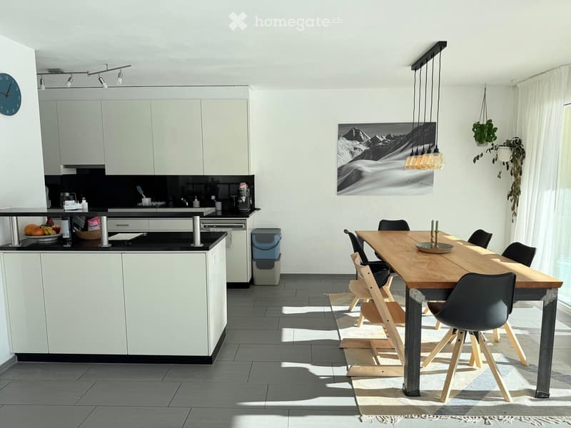 Modern kitchen with white cabinets, black countertops, a wooden dining table with chairs, pendant lights, and a large mountain-themed picture on the wall.