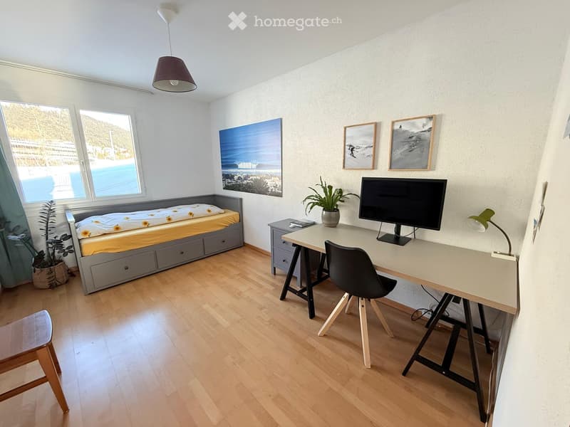 Cozy room with a bed, desk, chair, and a small stool. Features a large window with a view, a hanging lamp, and several framed pictures on the wall.