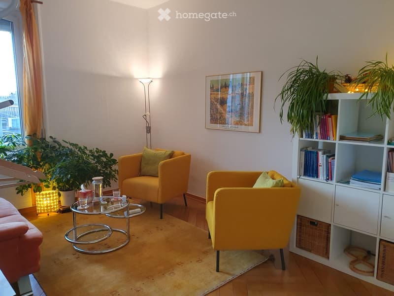 Bright living room with wooden flooring, a pink sofa, two yellow armchairs, a glass coffee table, a white bookshelf, and a floor lamp. A potted plant and a picture frame are also visible.