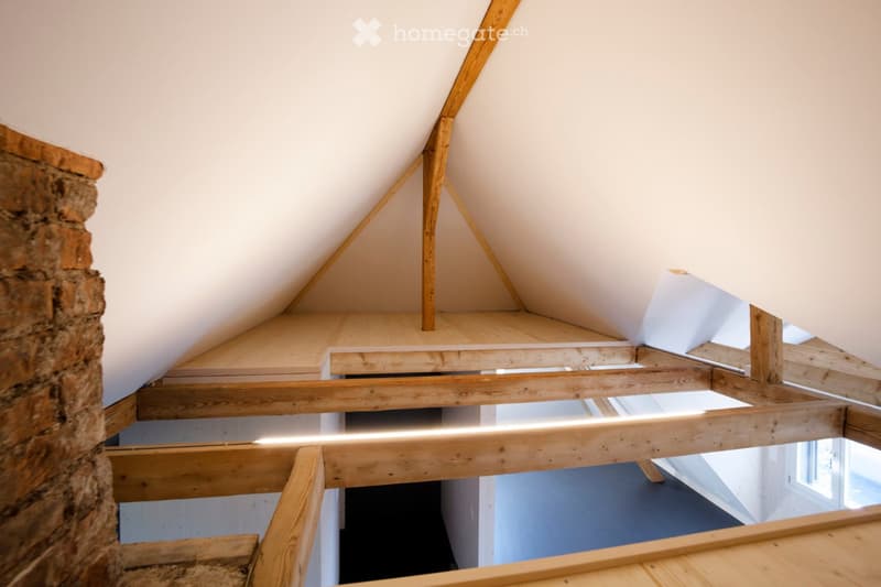 View of an attic or loft space with exposed wooden beams and a slanted ceiling. The area is unfinished, with visible wooden framing and a small window letting in natural light.
