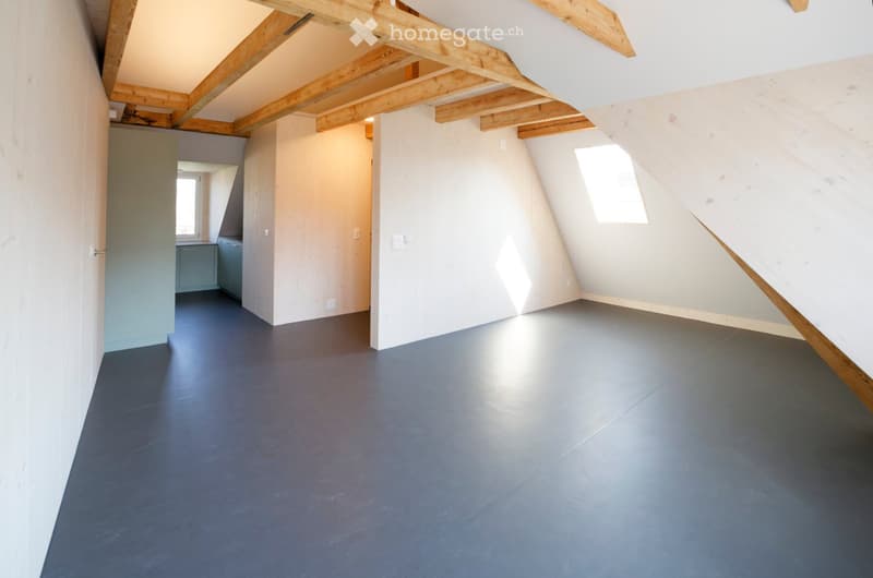 Empty attic room with exposed wooden beams, grey walls, dark grey floor, and two windows allowing natural light.