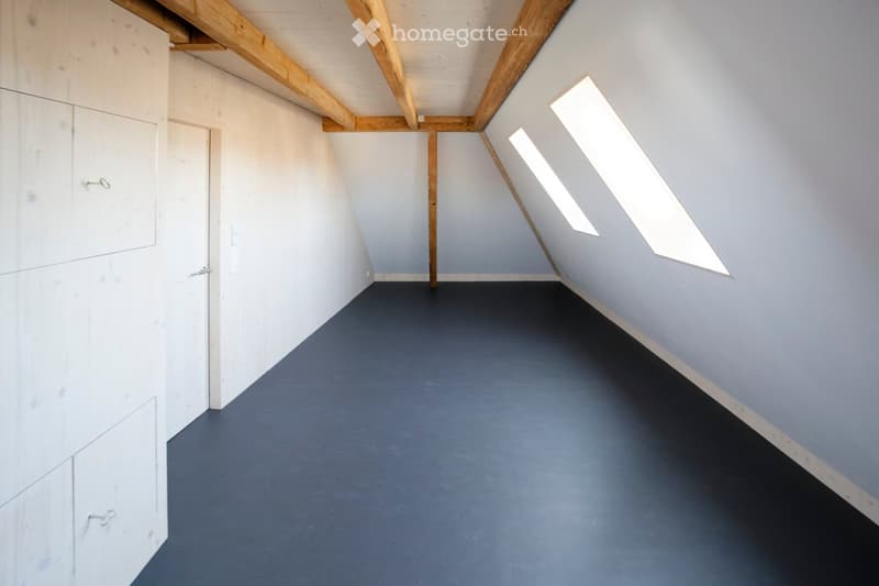 Empty attic room with exposed wooden beams, grey walls, and skylights allowing natural light. The floor is painted dark grey, and there is a white built-in cabinet on the left side.