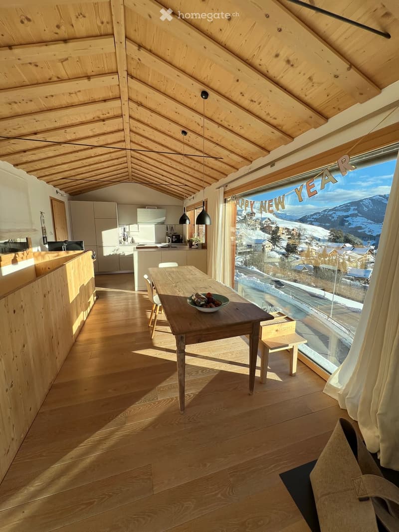 Bright, wooden-floored room with a dining table, chairs, a small kitchen area, and large windows offering a scenic mountain view.