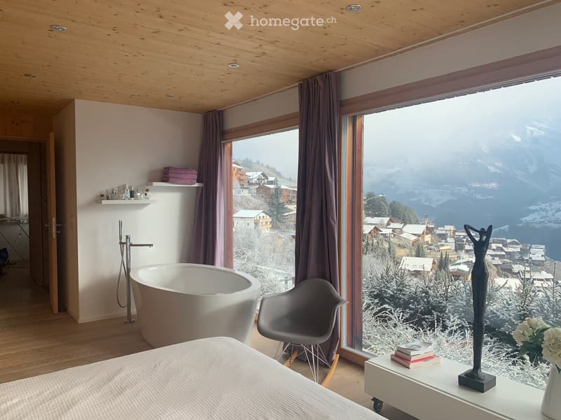 Modern bedroom with a large window offering a scenic mountain view, featuring a bed, a sleek chair, a bathtub, a shelf with towels, and a decorative statue on a table.