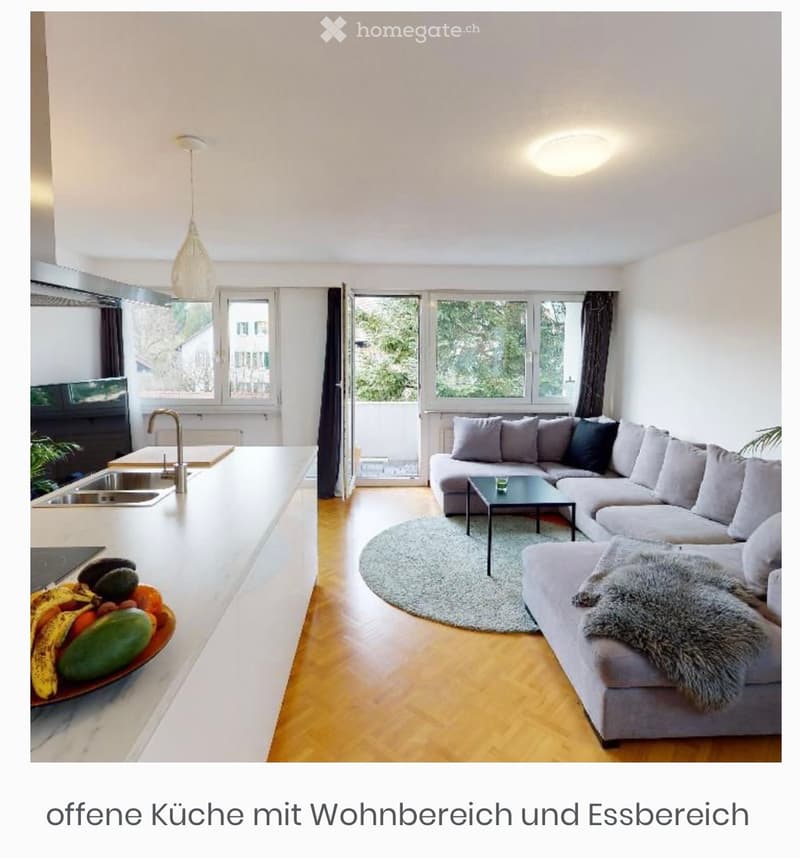 Open-plan living and dining area with a grey sectional sofa, coffee table, and a kitchen area featuring a sink, faucet, and a bowl of fruit on the counter.