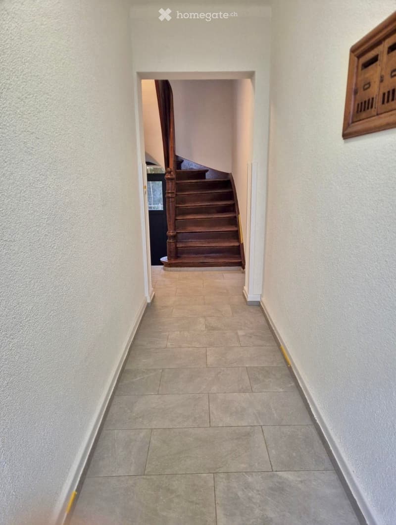 A narrow hallway with tiled flooring and light-colored walls, leading to a wooden staircase with a handrail.