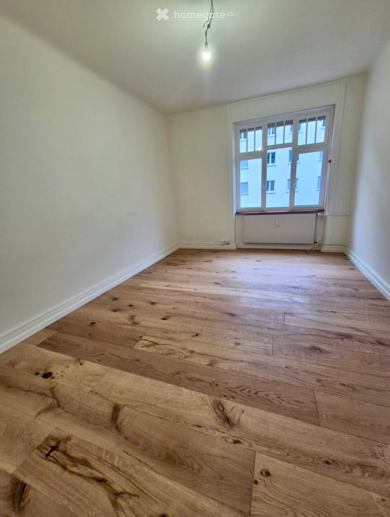 Empty room with wooden flooring, white walls, and a large window with a radiator below it.