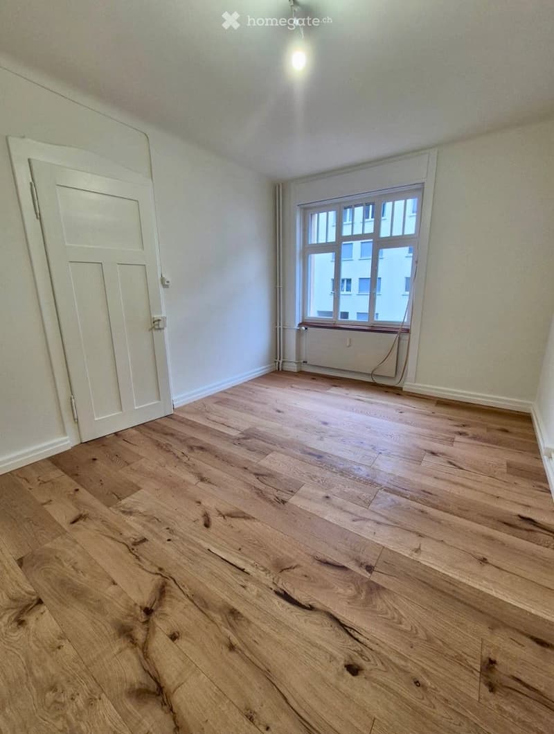 Empty room with wooden flooring, white walls, a single window, and a white door.