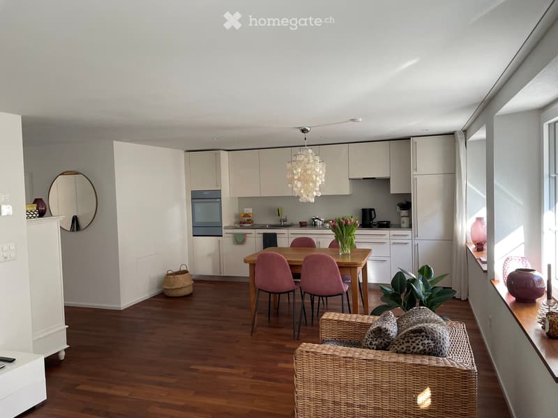 A modern, open-concept living and dining area with a kitchen in the background. The space features a wicker sofa with patterned cushions, a wooden dining table with pink chairs, a vase with flowers on the table, a round mirror on the wall, and a kitchen w