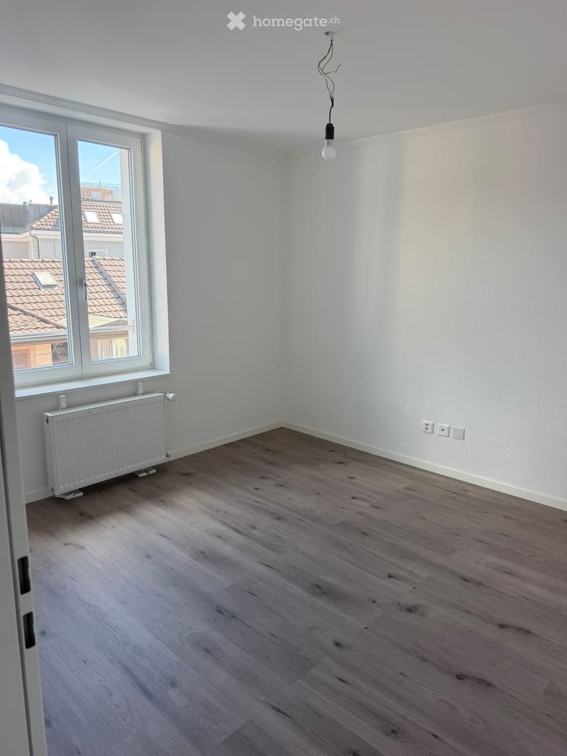 Empty room with light wooden flooring, white walls, a radiator, and a large window offering a view of rooftops.