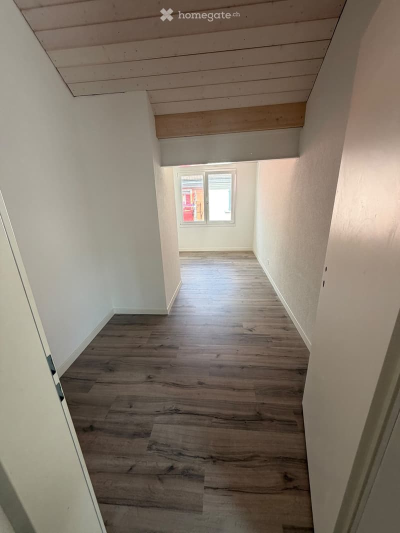 Empty hallway with wooden flooring and white walls, featuring a window at the end and a slanted wooden ceiling.